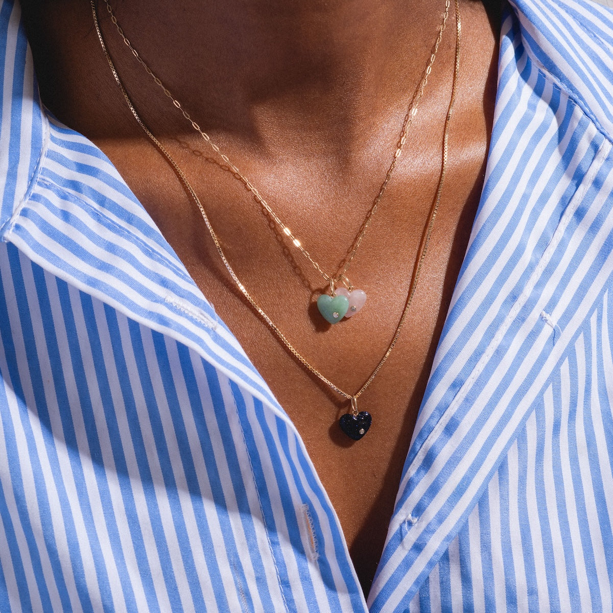 A person wears a blue and white striped shirt and three Solid Gold Full Heart Crystal Charms with green, pink, and dark blue crystals layered as necklaces on their chest.