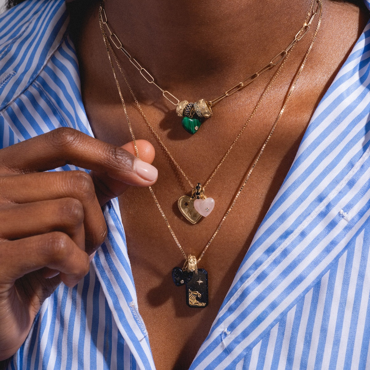 A person in a blue and white striped shirt displays three layered gold necklaces, each with a unique pendant: a green stone, the Solid Gold Full Heart Crystal Charm, and a black rectangular charm with gold detailing.
