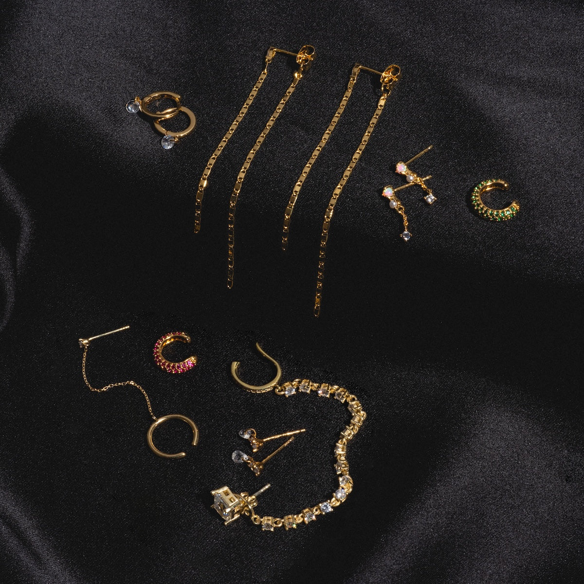 Ear Cuff with CZ Dangle Chain, displayed among assorted gold jewelry—hoop and drop earrings, a bracelet, and gemstone stud earrings—arranged on black satin fabric.