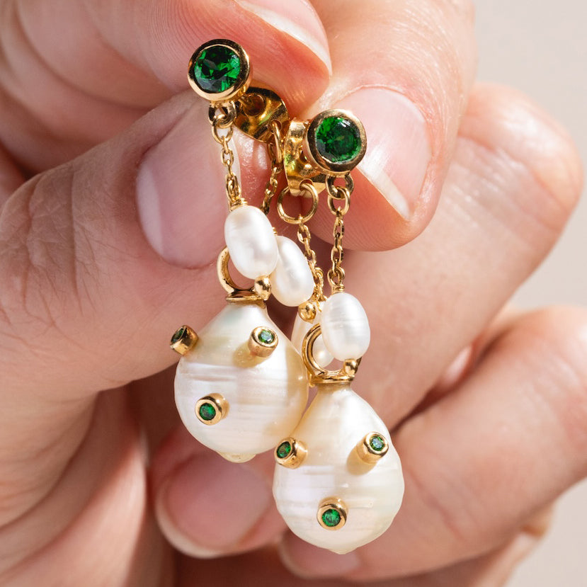 A hand holds the Baroque Pearl Droplet Earrings, featuring large baroque pearls accented with sparkling green CZ and gold studs, topped with smaller pearls and green stones in a 14k gold plated setting.