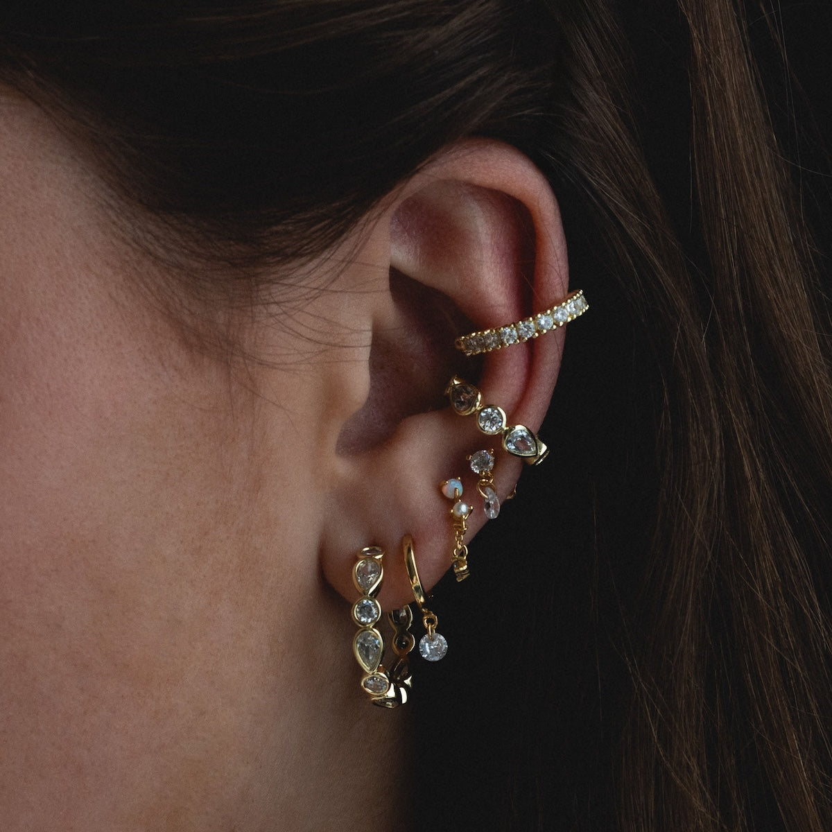 A close-up of a persons ear adorned with multiple gold earrings, featuring Isla Hoops and other elegant designs in 18k yellow gold with simulated diamonds, all set against a dark background.