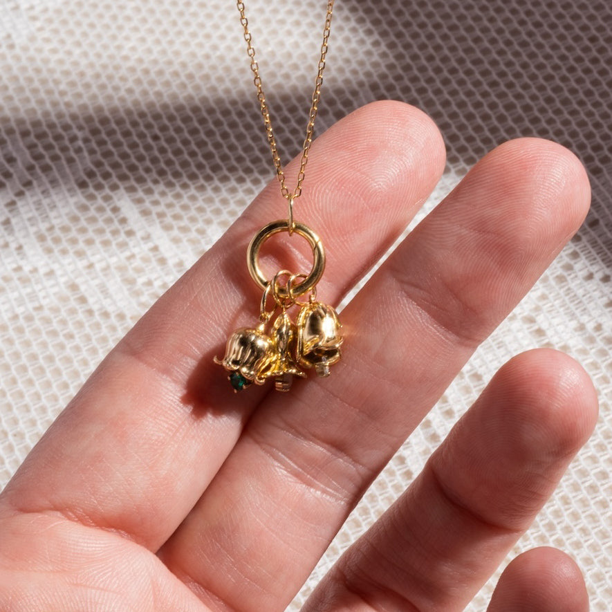 A hand displays the Solid Gold Birth Flower Bud Charm necklace, featuring a circular pendant and three small gold charms, including a birth flower charm and bead, set against textured white fabric—ideal for fans of personalized jewelry.