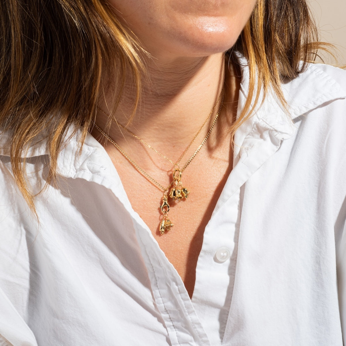 A woman in a white button-up shirt wears layered gold necklaces, including the Solid Gold Birth Flower Bud Charm; her brown highlighted hair is styled loose, with the photo cropped just below her nose.