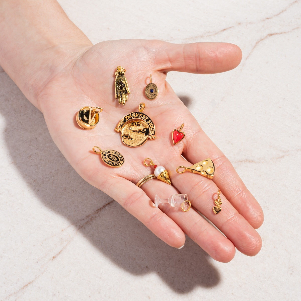 A hand displays Charms by Hunt of Hounds—gold vermeil pieces like a pizza slice, red heart, hand, round face, and zodiac coin—on a light marble surface.