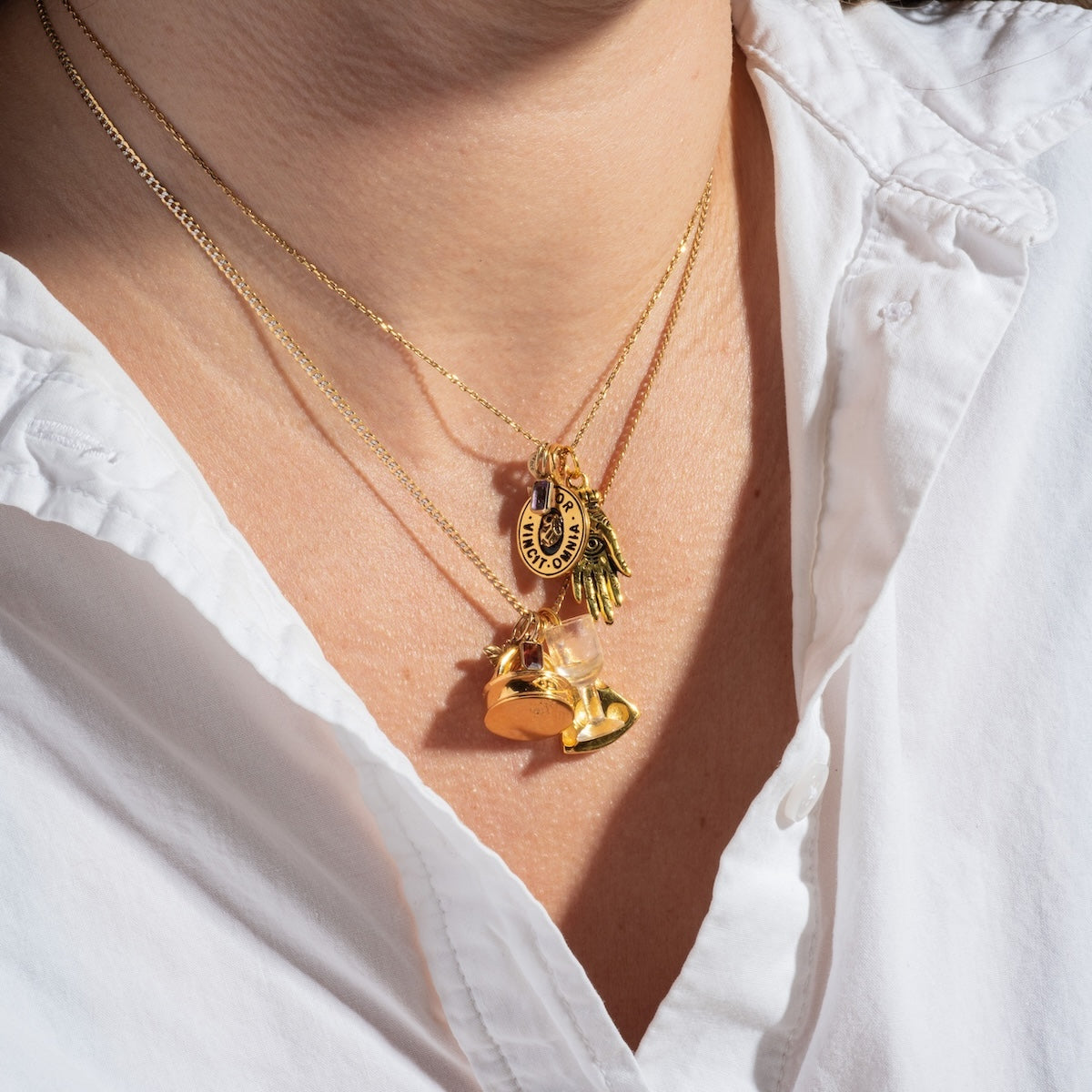 A person wearing a white shirt and three gold vermeil Charms by Hunt of Hounds necklaces, each featuring unique pendants: a heart, a padlock, a key, and a circular charm.