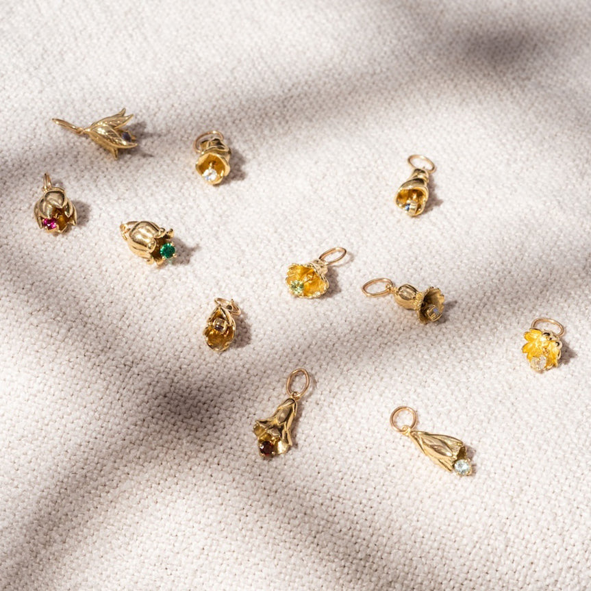 Solid Gold Birth Flower Bud Charms with colorful gemstones, shaped like flowers and buds, are scattered across textured beige fabric with soft shadows in the scene.