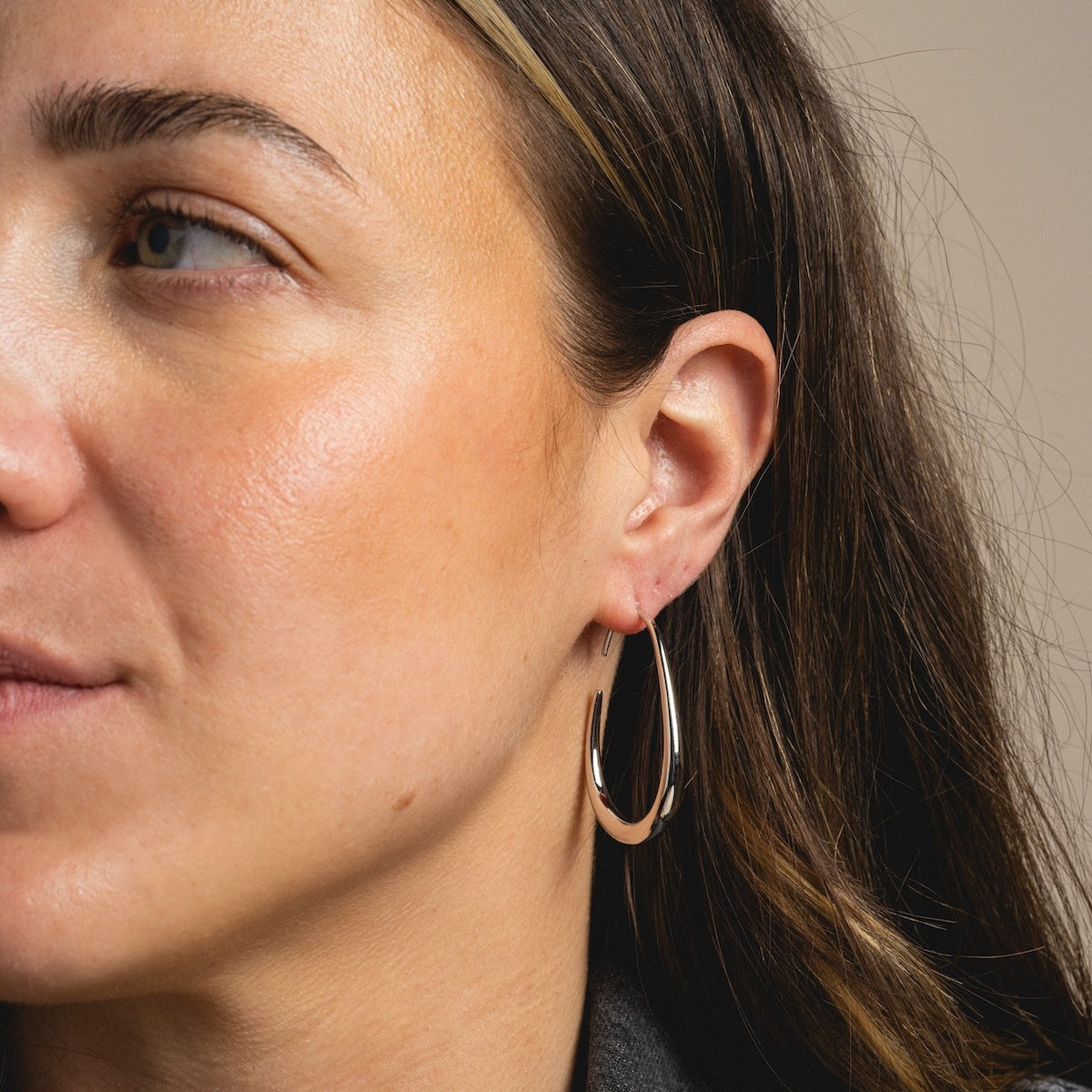 A close-up of a woman with long brown hair and clear skin, facing slightly left, highlights her ear adorned with a large, shiny teardrop-shaped Fluid Hoop Earring.