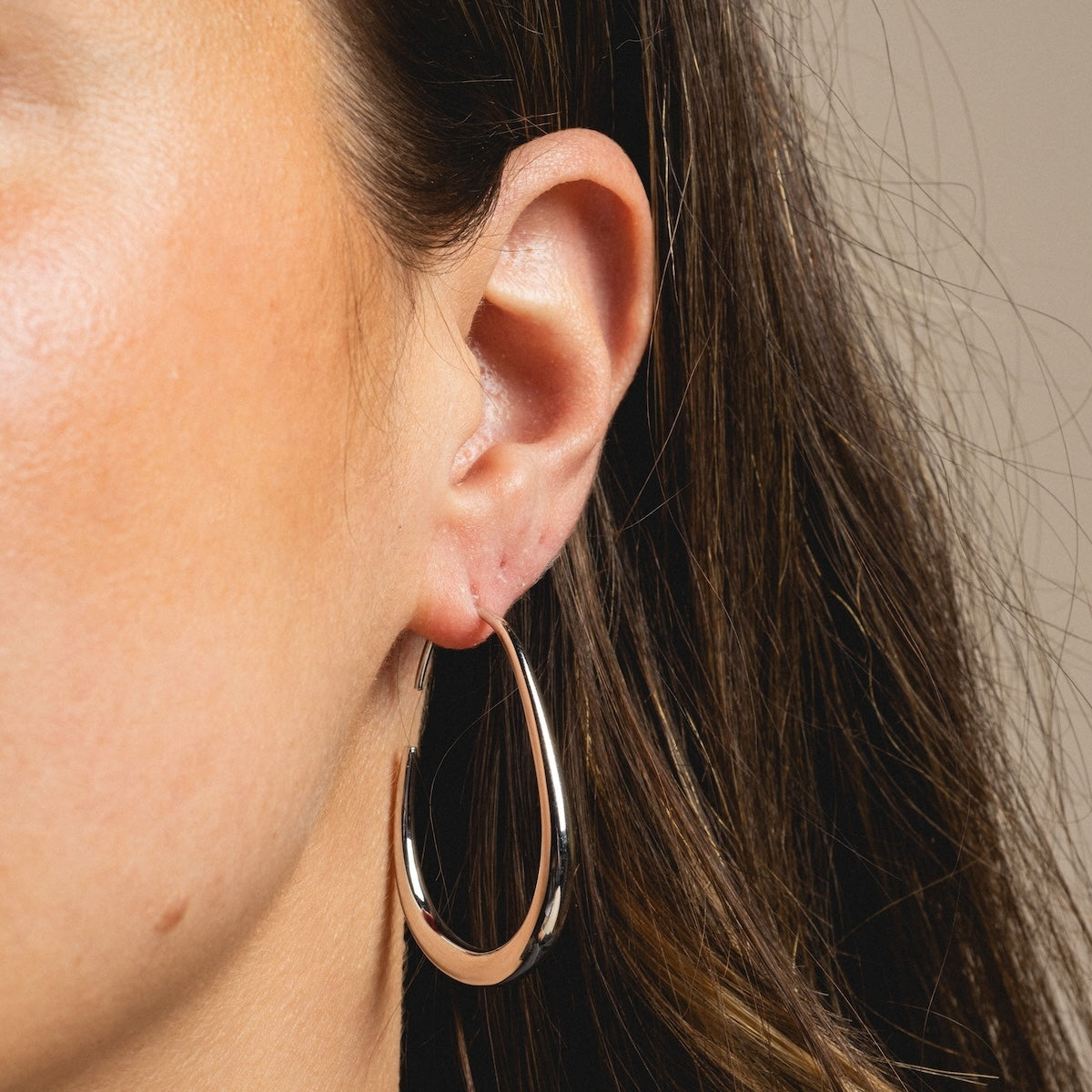 A close-up shows a womans ear with long brown hair, wearing Fluid Hoop Earrings in a shiny yellow gold plated finish. The softly lit, neutral background highlights the thick and elegant design.