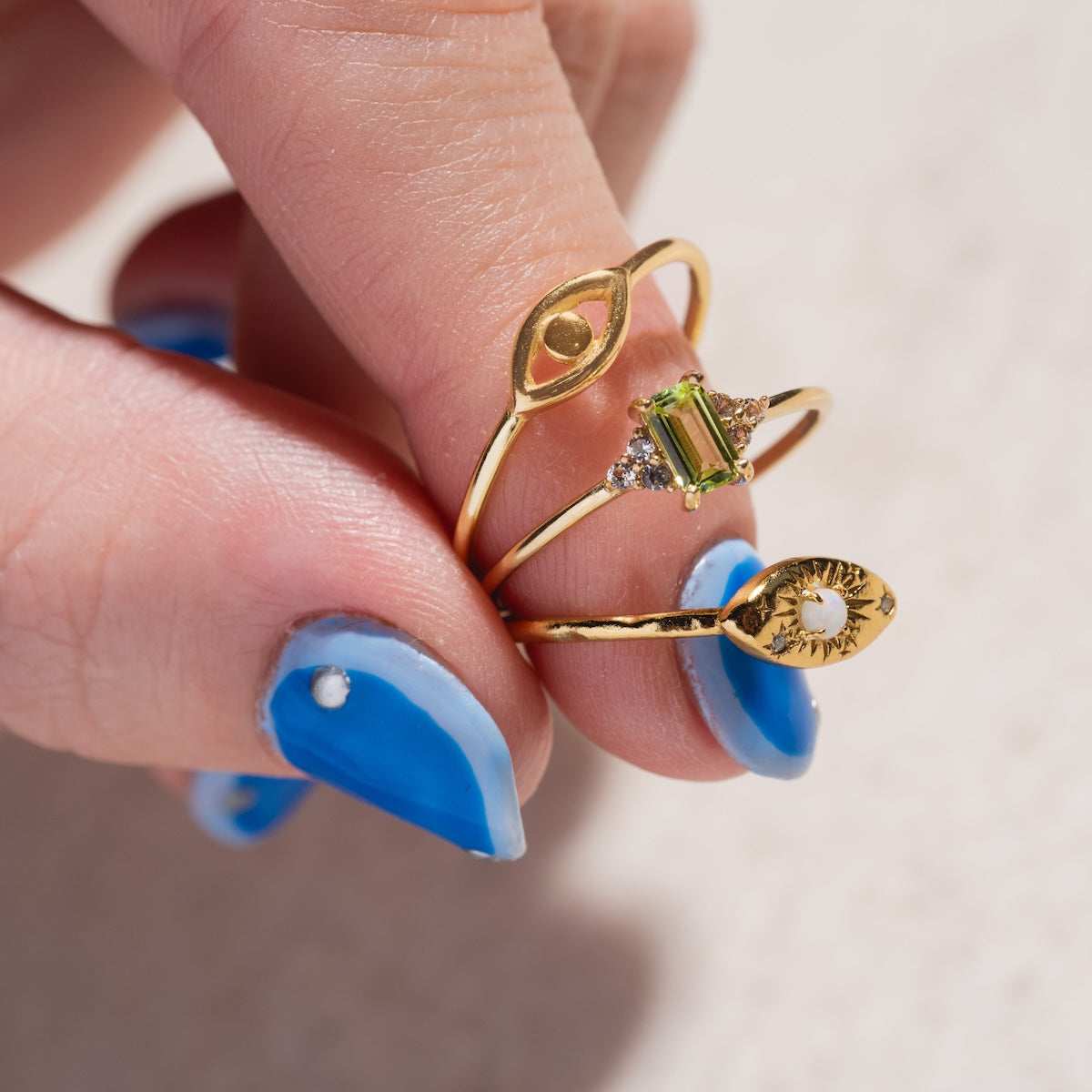 A hand with blue and white nail art holds three gold rings: the Opal Amulet Ring, one featuring a green gemstone, and another adorned with a white pearl—each displaying unique design details and elegant style.