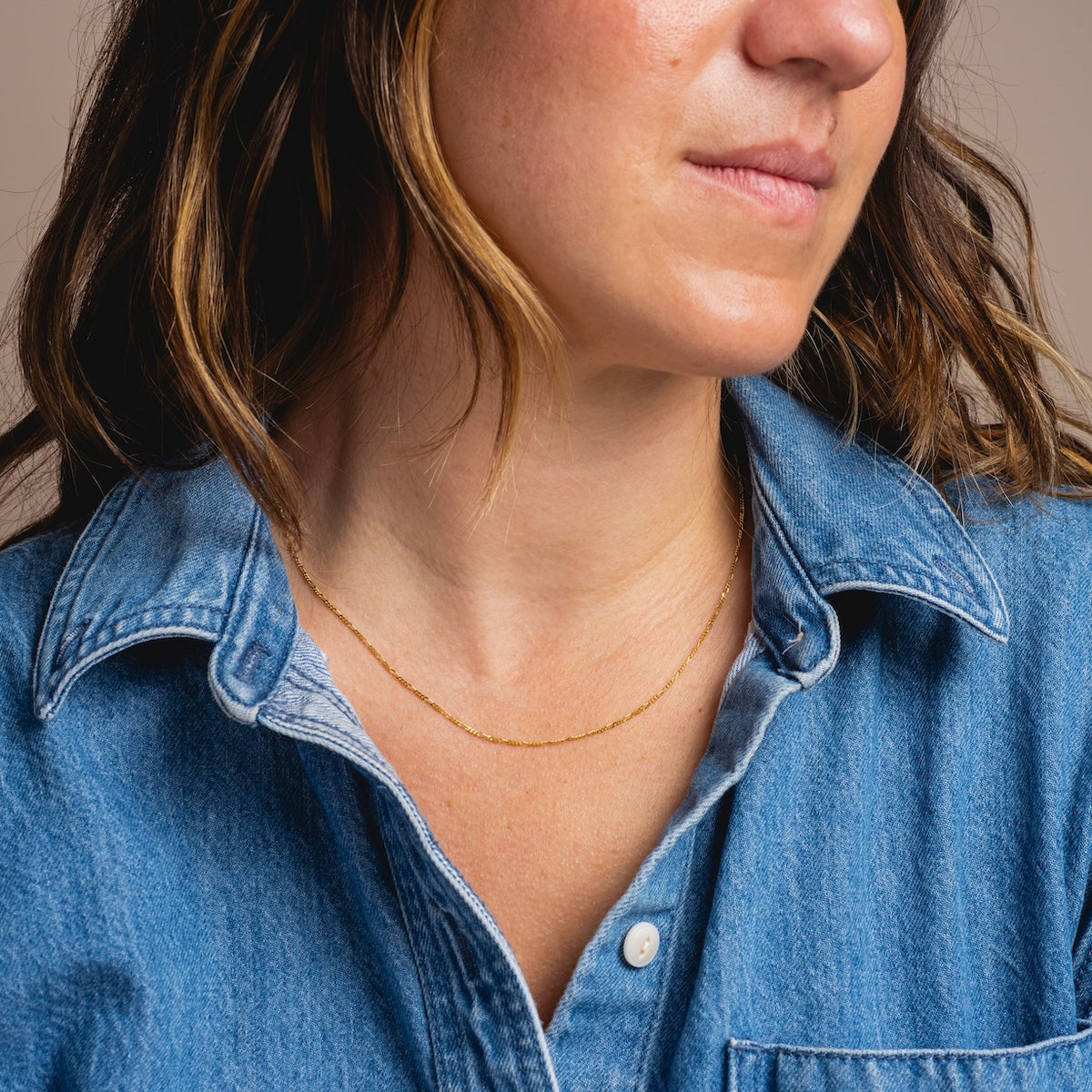 A woman with light brown wavy hair and a neutral expression wears a blue denim shirt and the Solid Gold Figaro Chain necklace, shown from the shoulders up against a plain background.
