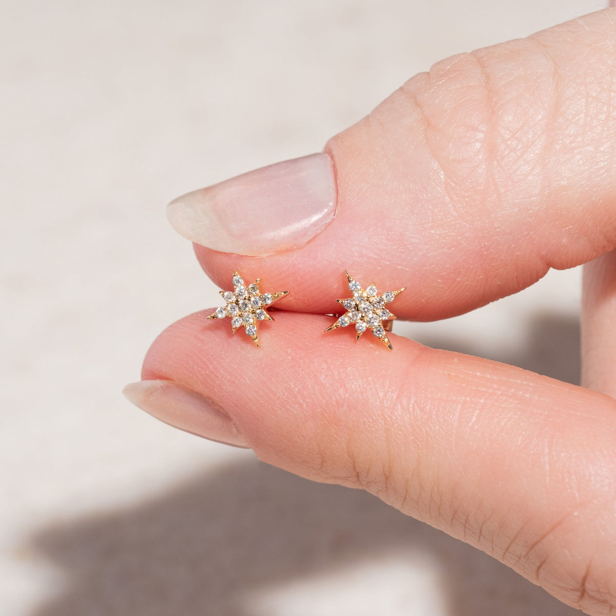 A hand holds the Solid Gold Diamond Starlight Studs—two small earrings shaped like eight-pointed stars, each sparkling with diamonds—highlighting their elegant, celestial design against a light neutral background.