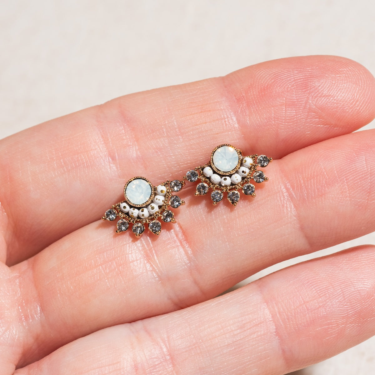 A close-up of a hand holding the Nico Fan Earrings in White Opal, featuring an ornate gold-plated design with an oval opalescent center, surrounded by small white and metallic stones in a fan shape.