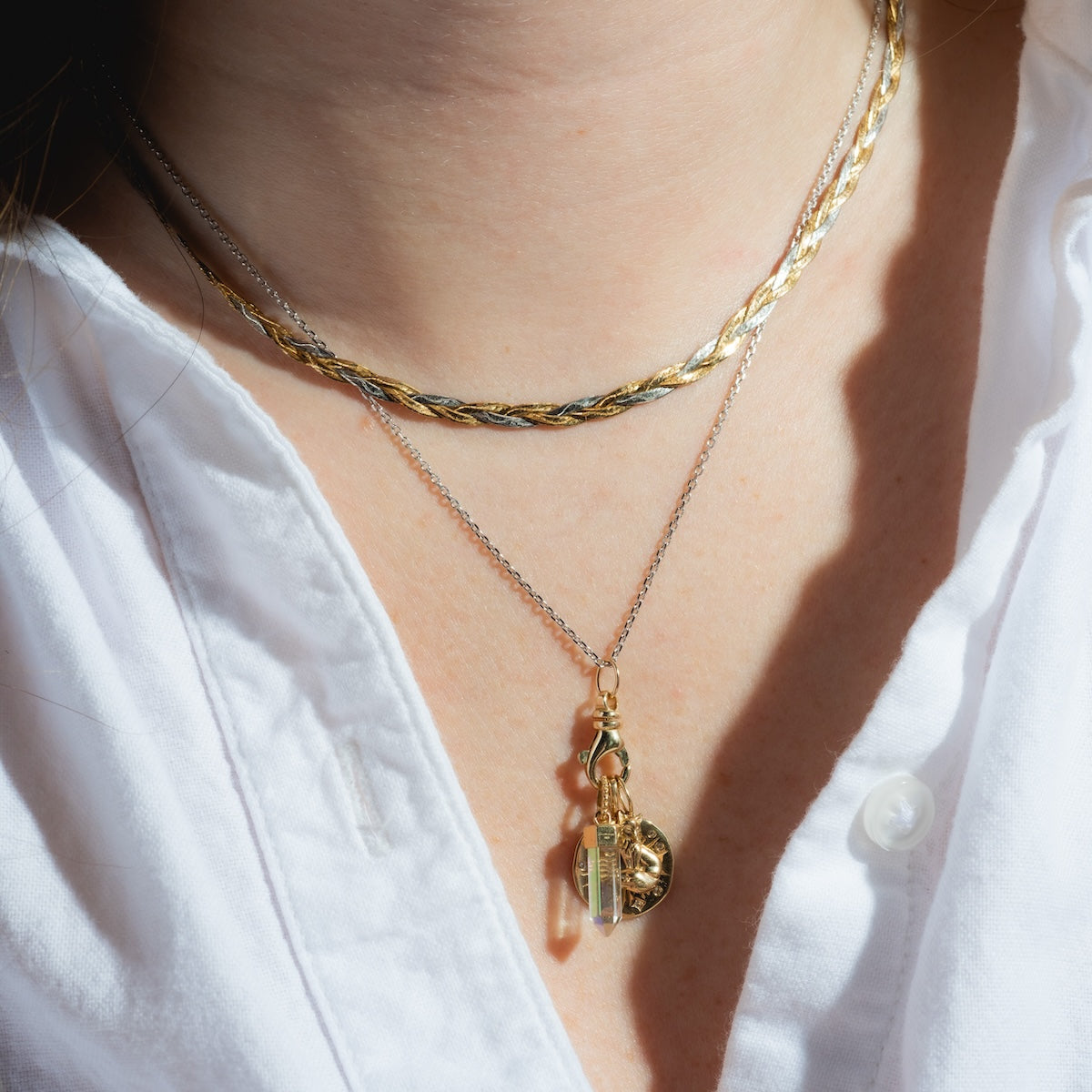 A close-up of a person wearing a white button-down and layered gold necklaces—one is the Solid Gold Simple Glimmer Gold Chain, paired with styles featuring a round coin pendant and a rectangular crystal.