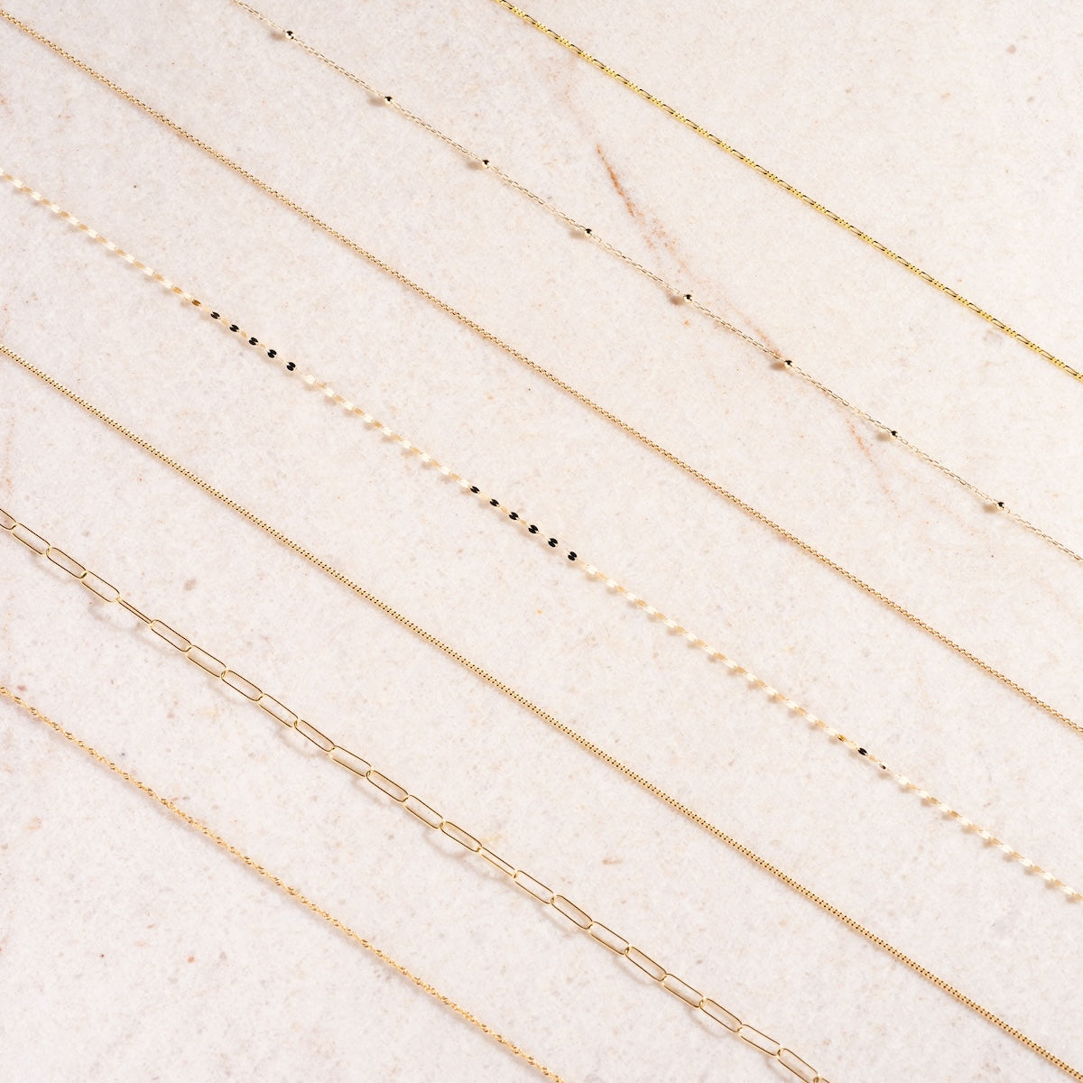 Nine delicate necklaces, including the Solid Gold Chain for Charms, are arranged parallel on a marble-like surface, displaying styles such as simple chains, dainty beads, and rectangular links.