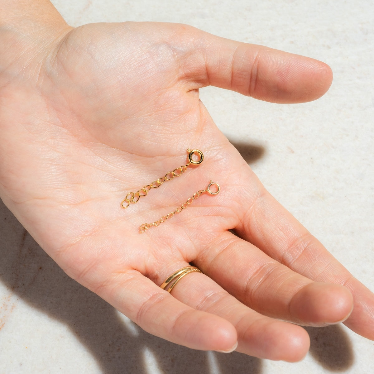 A hand with two gold rings holds two short 14k gold plated Necklace and Bracelet Extenders with clasps, ideal for adjusting jewelry length, shown against a light background.