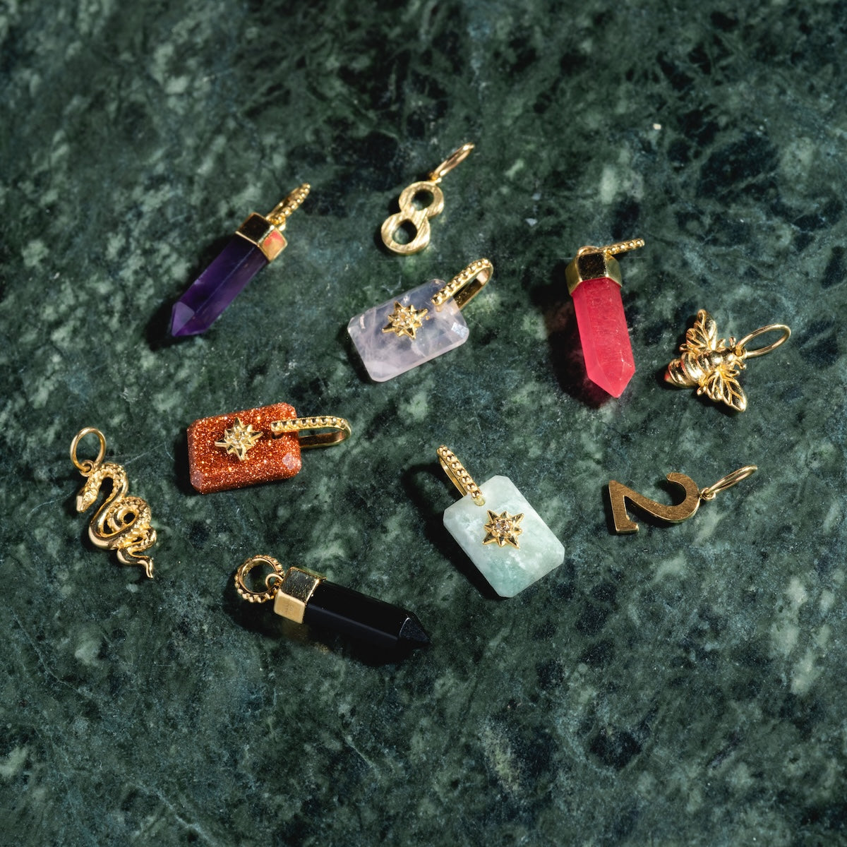 The Solid Gold Crystal Amulet Charm for Manifestation, along with crystal points, number charms, and star and animal shapes, is displayed on a dark green marble surface.