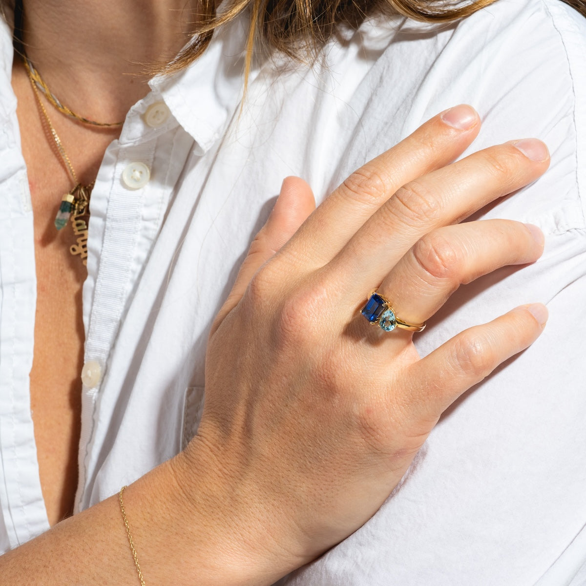 Wearing a white button-up shirt, a person shows off their hand adorned with the Solid Gold Toi et Moi Colorful Gemstone Ring, complemented by gold necklaces and a delicate gold bracelet as sunlight highlights their jewelry and skin.