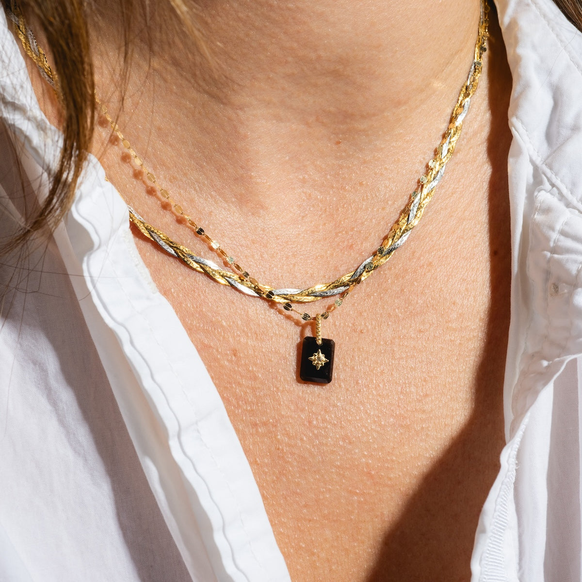 A person wearing a white shirt and layered gold necklaces, including the Solid Gold Crystal Amulet Charm for Manifestation, which features a black rectangular pendant with a small star design.