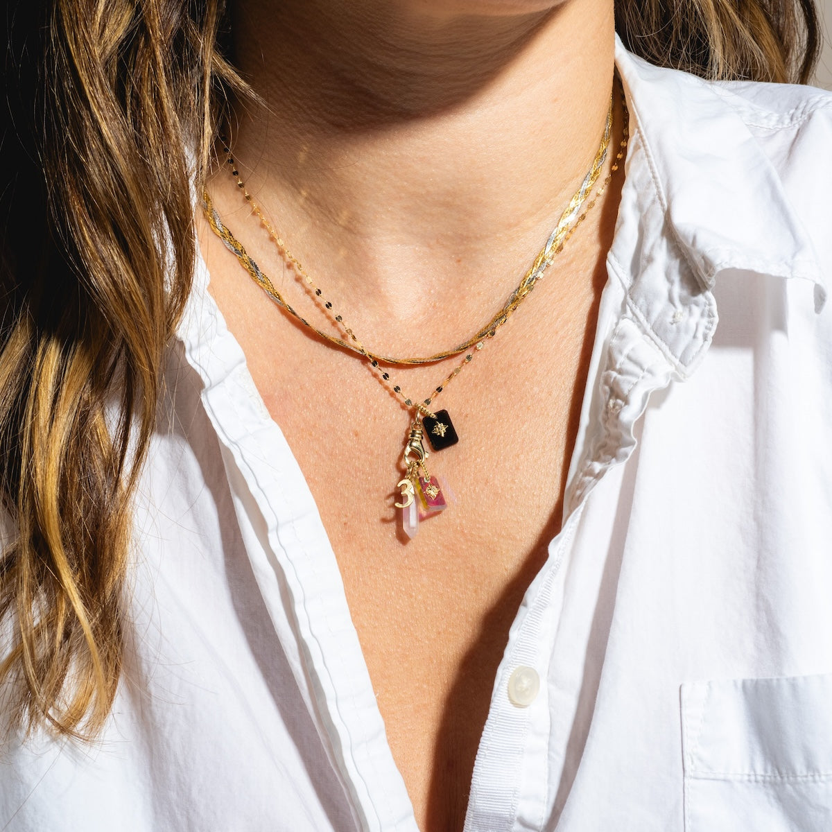 A person wears a white button-up shirt and layered gold necklaces, including the Solid Gold Crystal Amulet Charm for Manifestation decorated with colorful pendants. Only their neck and part of their chest are shown.