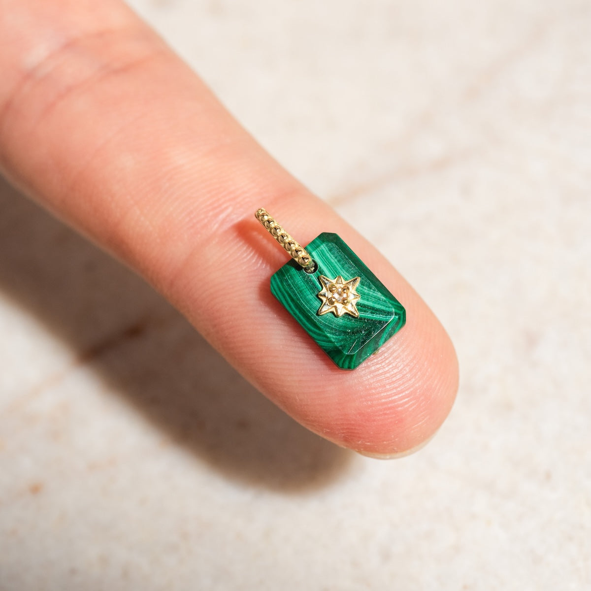 The Solid Gold Crystal Amulet Charm for Manifestation, a small green rectangular pendant with a gold star design, is shown resting on a fingertip against a light background to highlight its size and detail.