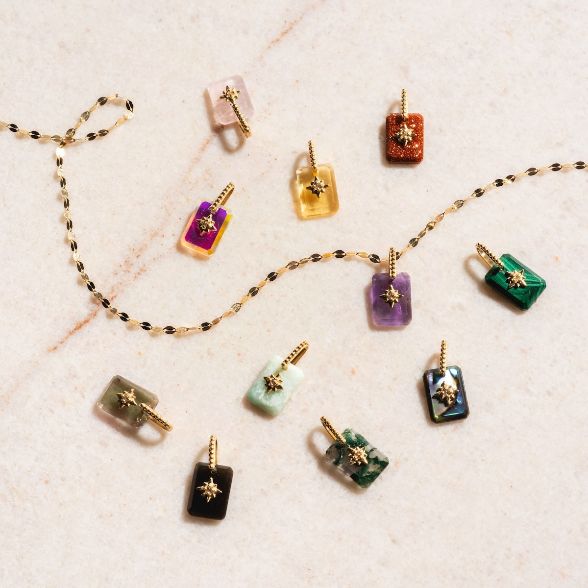 The Solid Gold Crystal Amulet Charm for Manifestation features rectangular natural crystal pendants with gold star accents in green, purple, black, and pink, displayed on marble beside a twisted gold chain.