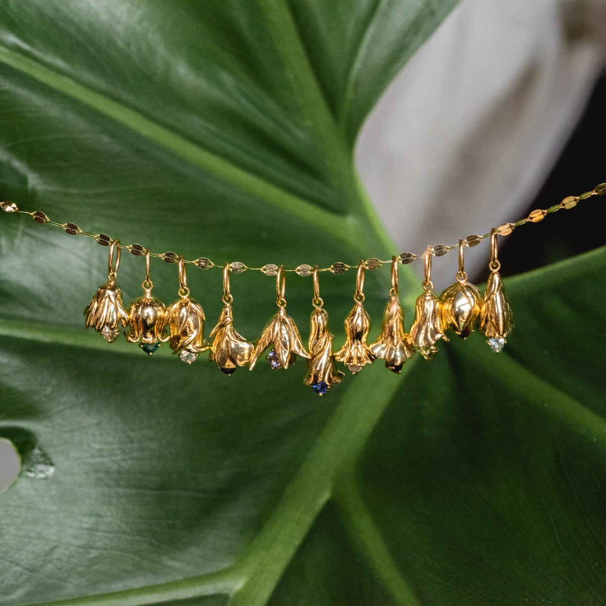 The Solid Gold Birth Flower Bud Charm, featuring a delicate flower-shaped pendant and sparkling gemstones, is elegantly draped on a large green tropical leaf. Each charm offers unique personalized jewelry details.