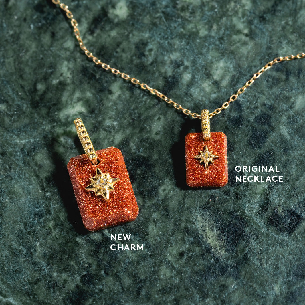 A gold manifestation necklace with a glittery starburst pendant rests beside the Solid Gold Crystal Amulet Charm for Manifestation on a green stone surface. Labels read ORIGINAL NECKLACE and NEW CHARM.