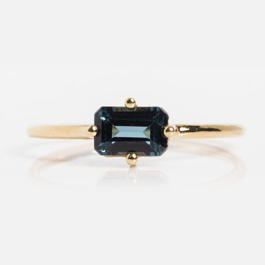The 14k Mod Love Birthstone Ring showcases a yellow gold band with an emerald-cut deep blue gemstone in a minimalist prong setting, displayed against a white background.