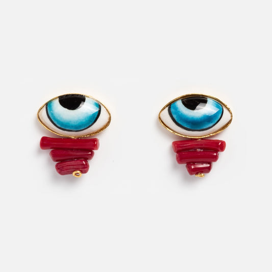 The Adira Blue Porcelain Evil Eye Stud Earrings feature blue eye-shaped studs with 21k yellow gold plating, hand-painted porcelain, and red coral-like bead clusters hanging below, all set against a plain white background.