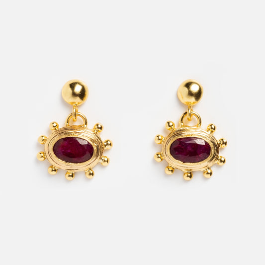 The Zand Ruby Drop Earrings feature small gold studs with dangling oval red stones, each set in a sunburst design and accented with gold beads—bold, bohemian-style earrings displayed on a plain white background.