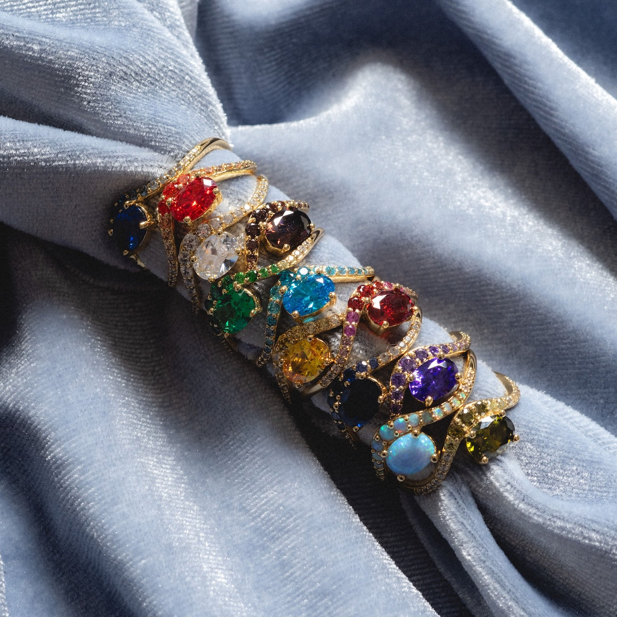 The Main Character Floating Ombre Birthstone Ring dazzles with red, blue, green, yellow, and purple stones displayed on soft light blue velvet fabric.