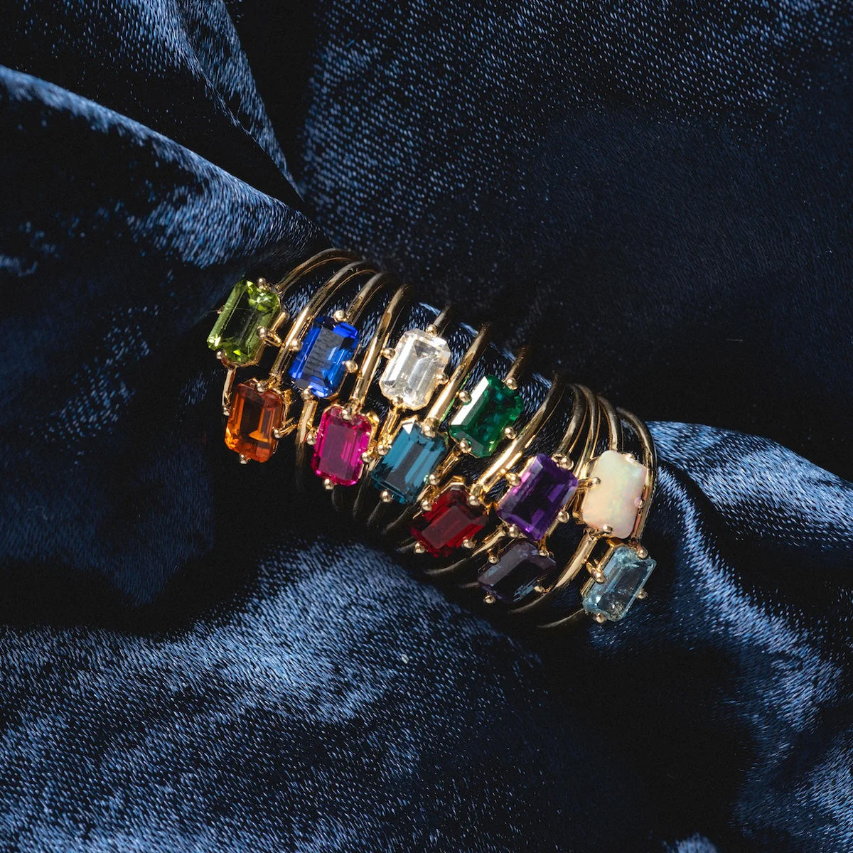 A 14k Mod Love Birthstone Ring, featuring a colorful rectangular gemstone, is displayed on dark blue textured fabric.