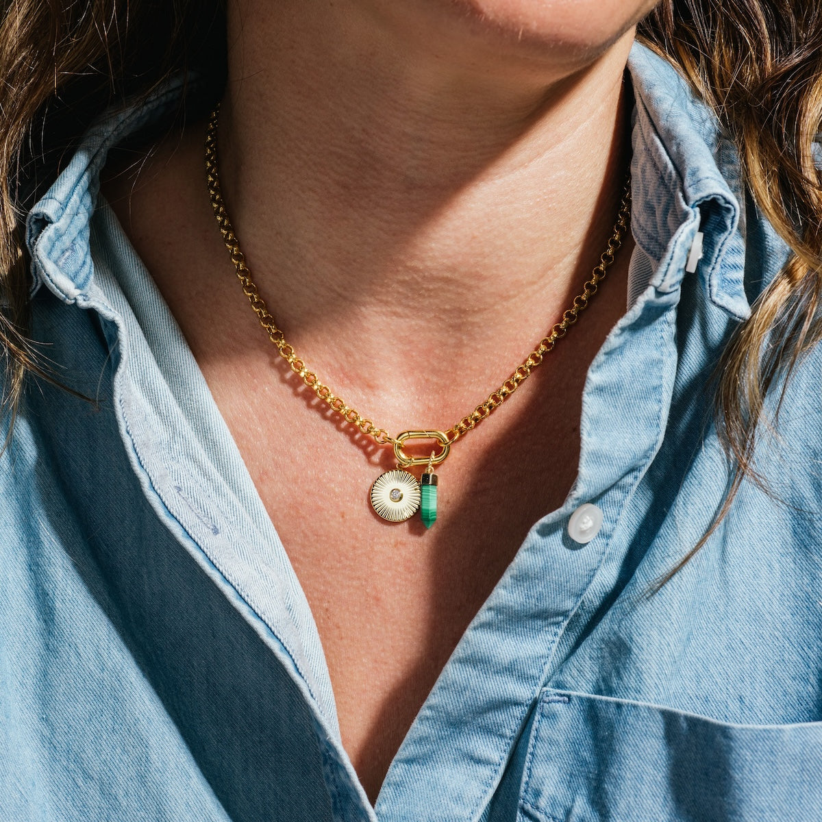 A person wears the Gold Filled Charm Holder Necklace by Local Eclectic, featuring two pendants—one circular and one turquoise—over a blue denim button-up. The 18k yellow gold filled chain brings added elegance to the outfit.