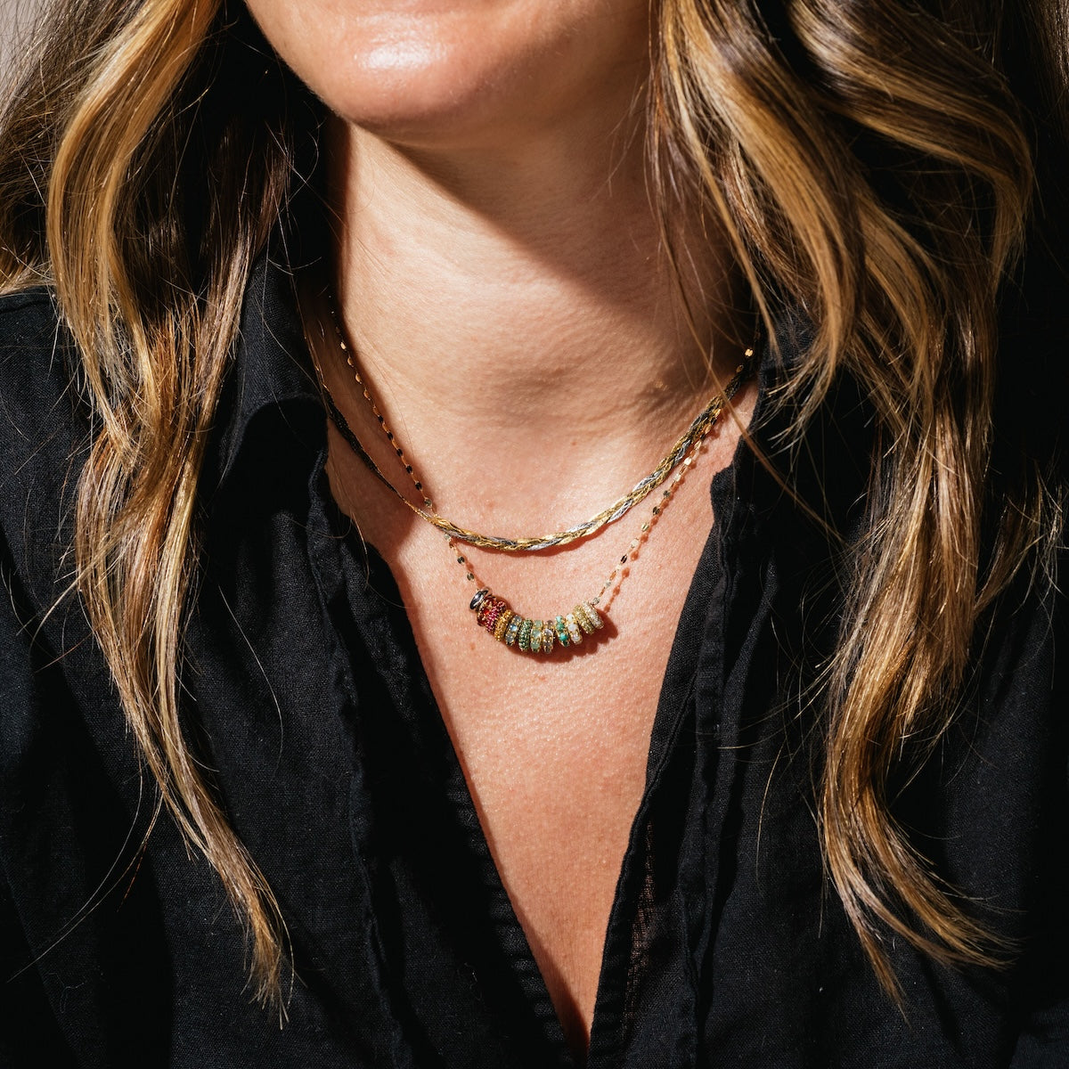 A woman with wavy light brown hair layers two gold necklaces; one features the Solid Gold Anniversaire Charm, adding a touch of milestone jewelry to her black shirt look.