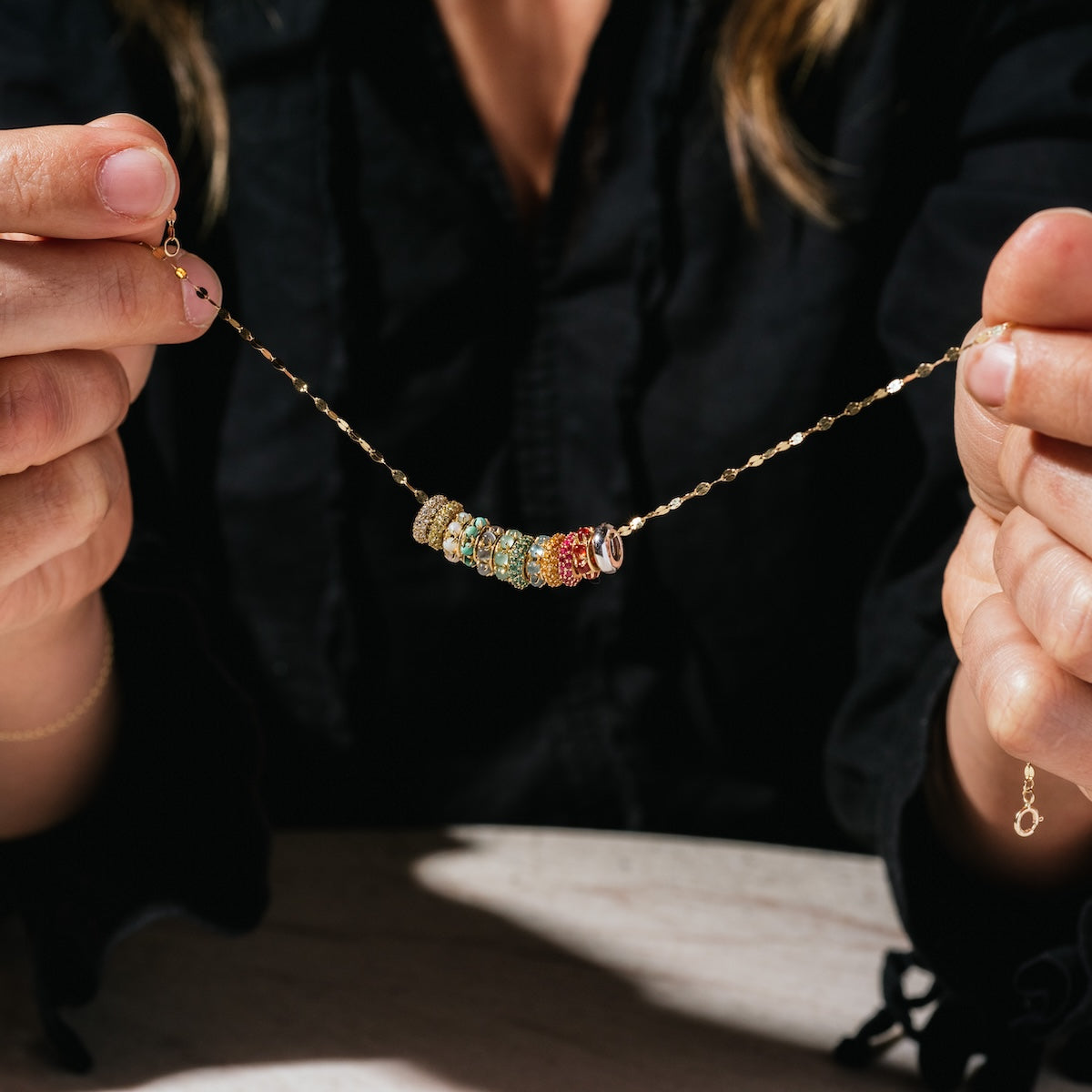 A person in a black shirt holds a gold chain necklace with colorful gemstone rings and Solid Gold Anniversaire Charms, showcasing this milestone jewelry on a light surface.