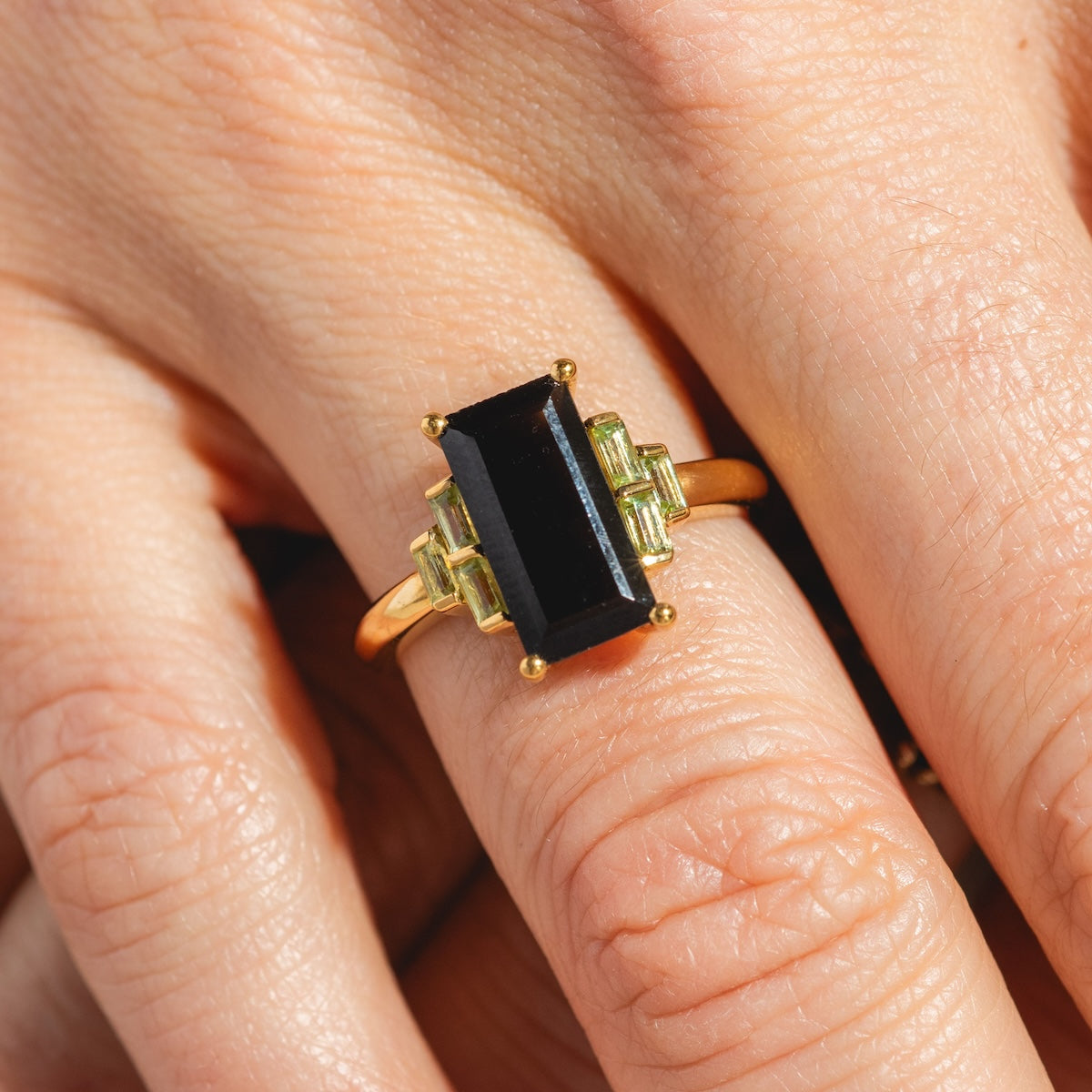 A hand wears the Bold Baguette Statement Ring—14k yellow gold plated, featuring a large black onyx center and small light green stones on each side.
