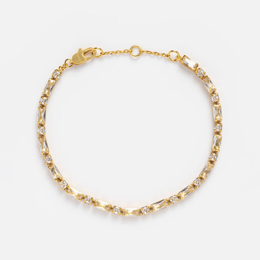 The Baguette Tennis Bracelet features alternating rectangular and round white CZ stones, 14k yellow gold plating, a lobster clasp, and an adjustable chain, shown on a white background.