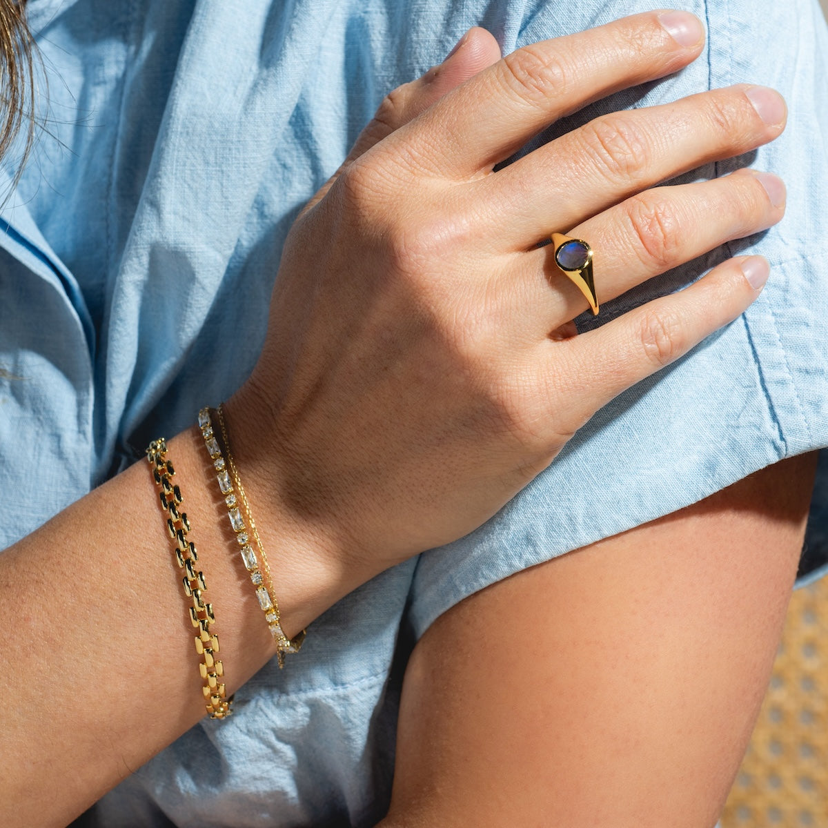 A person in a light blue shirt rests their hand on their arm, showcasing the Baguette Tennis Bracelet in 14k yellow gold plating and a gold ring with a dark stone.