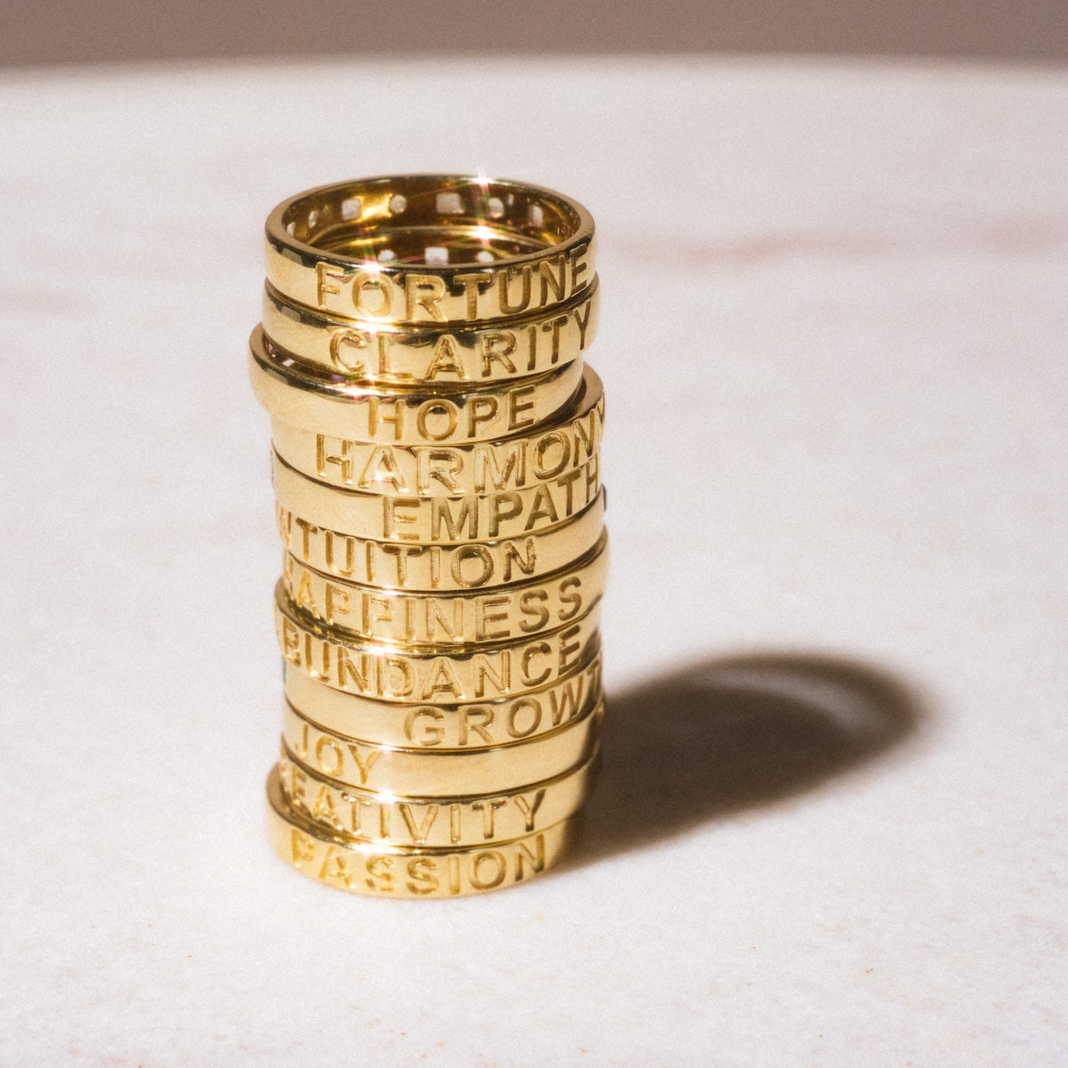 A stack of Solid Gold Within You Birthstone Band Sample Size 7 rings in 9k yellow gold, each 3.1mm wide and engraved with words like Fortune, Clarity, and Hope, forms an elegant and meaningful statement on a light surface.