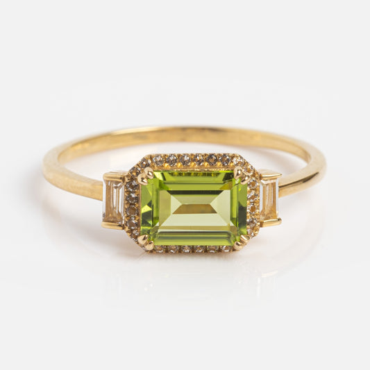 The Solid Gold Vintage Inspired East West Peridot Ring showcases a rectangular green gemstone at the center, accented by small clear stones and two baguette-cut clear stones on each side, radiating timeless elegance.