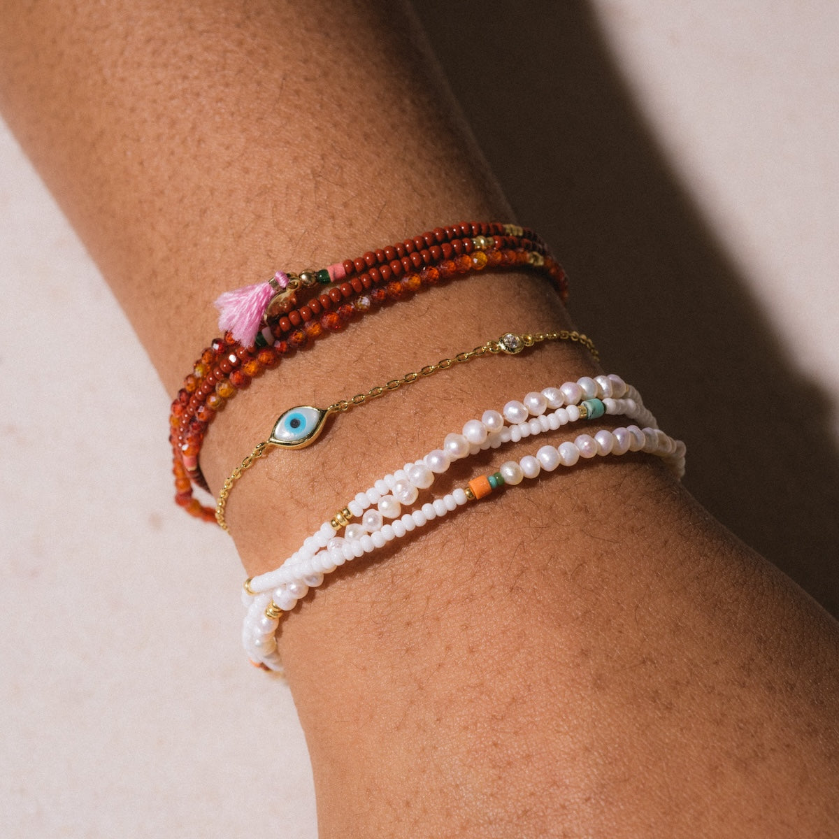 A close-up of a wrist with four bracelets: two red-orange beaded, the Madison Evil Eye Bracelet featuring a blue charm in 14k vermeil, and two white beaded bracelets with colorful accents—an elegant protective jewelry style.
