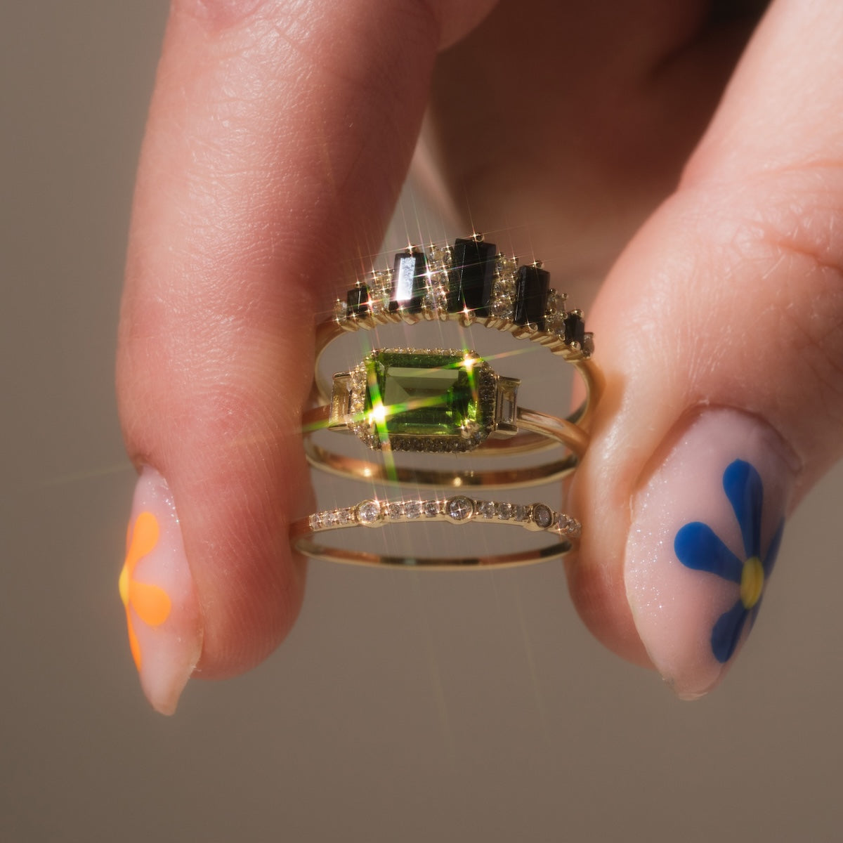 A hand with nails adorned in colorful flowers holds four gold rings, including the Solid Gold Delicate White Sapphire Stacking Band, each set with unique black, green, and clear stones.