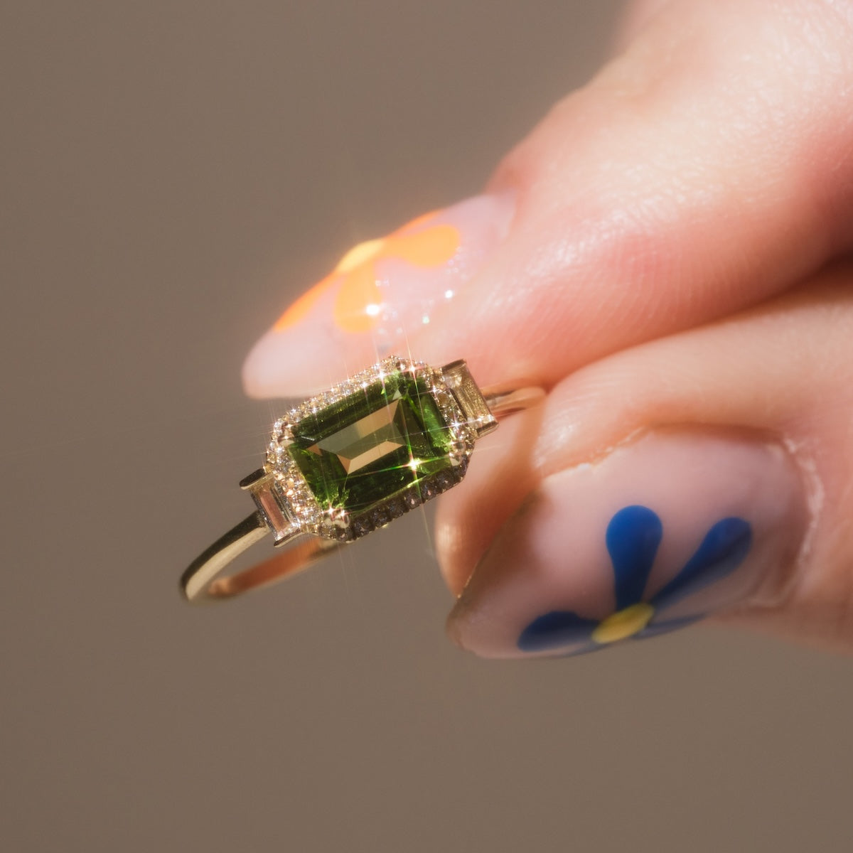 A hand with colorful floral nail art holds the Solid Gold Vintage Inspired East West Peridot Ring, featuring a rectangular green gemstone accented by small diamonds—a true vintage charm piece.