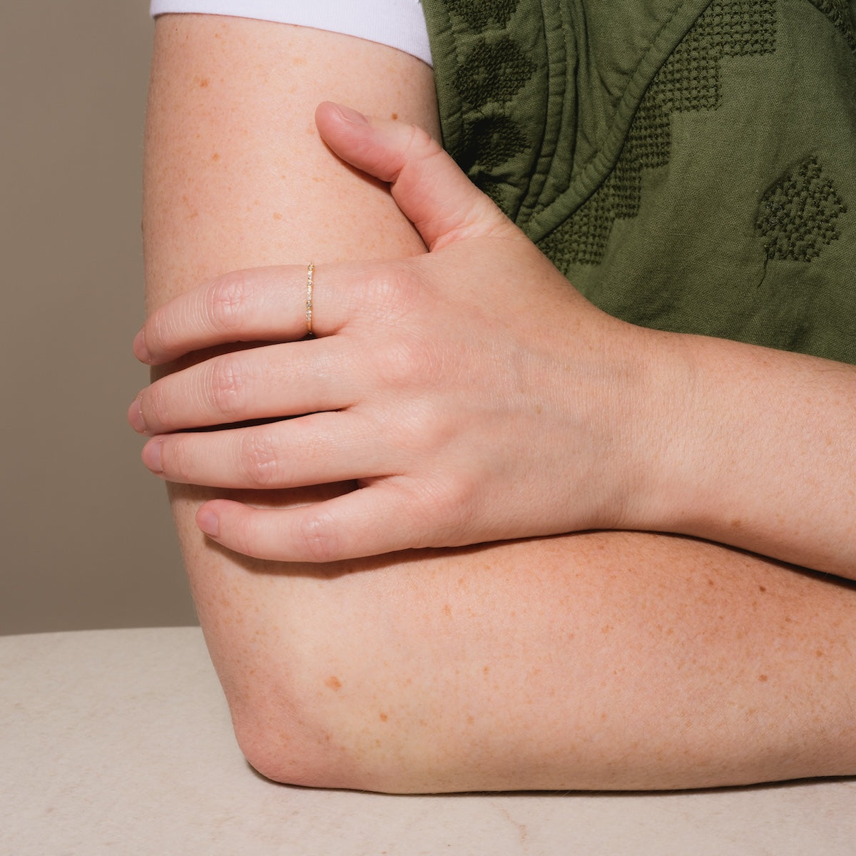 A person in a green textured top rests their left arm on a surface, gently holding it with their right hand, which wears the Solid Gold Delicate White Sapphire Stacking Band on the ring finger.