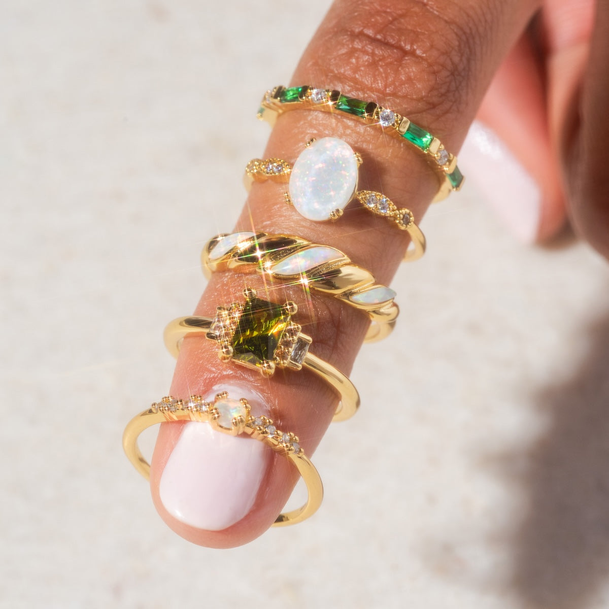 A hand with light pink nail polish displays five gold rings, including the Opal Sparkle Ring and a cubic zirconia ring with green stones, elegantly showcased on one finger against a light background.
