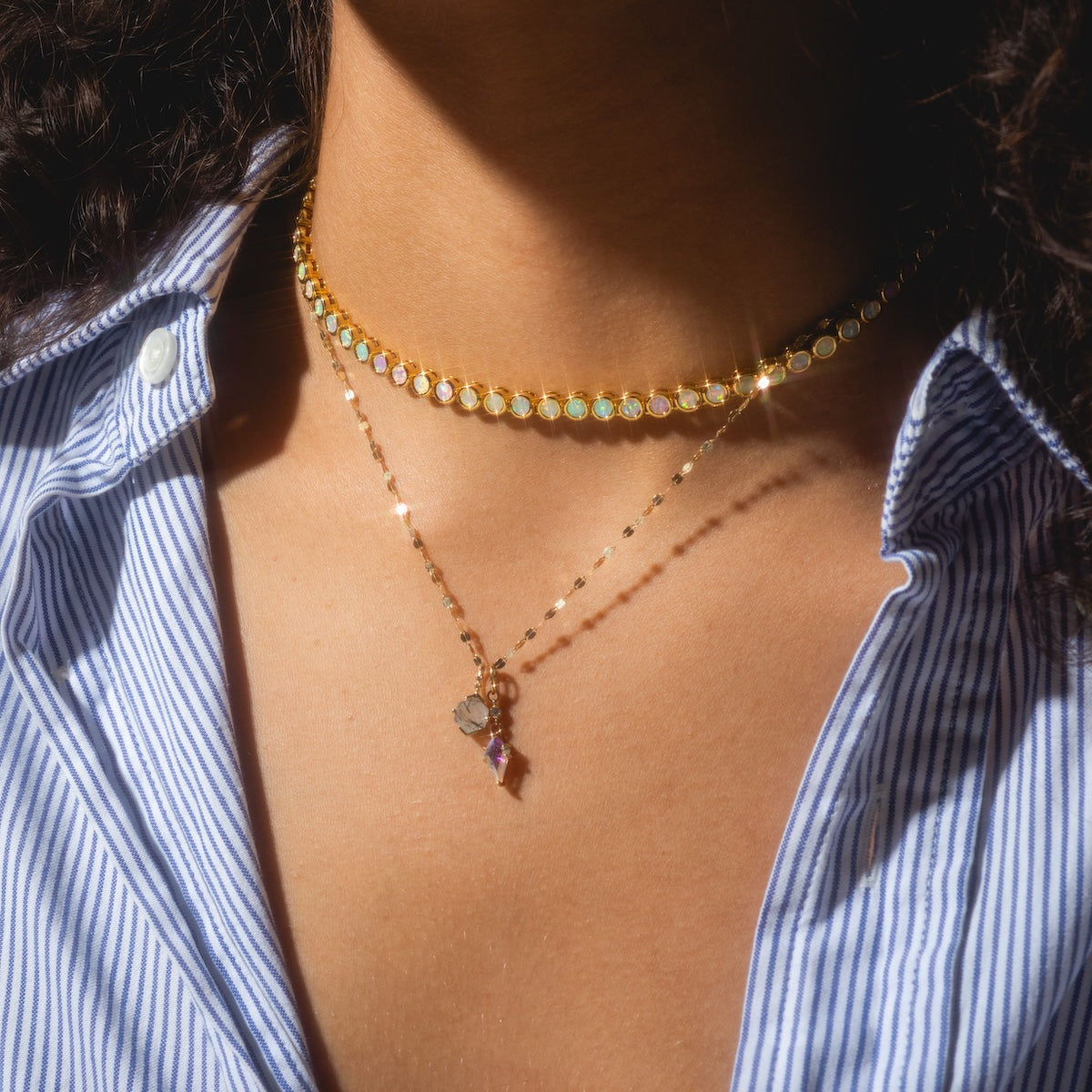 A person in a blue and white striped shirt wears two gold necklaces: an 18k gold plated choker with the Opal Ombre Tennis Necklace’s colorful stones and a longer chain with a heart pendant and small purple charm.