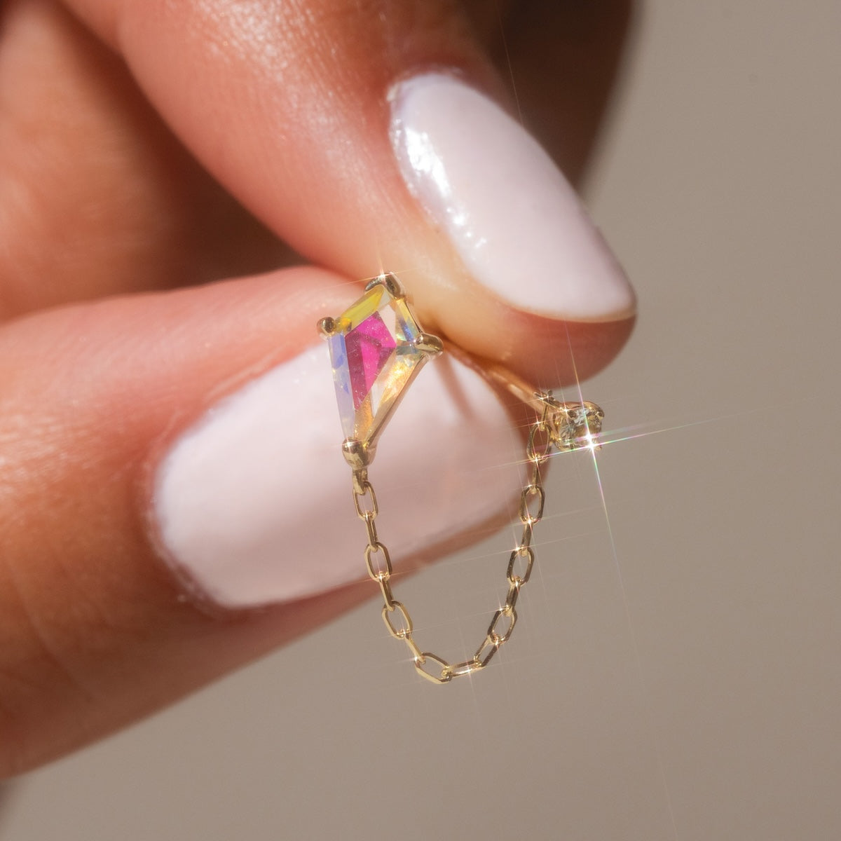 A hand with pale pink nail polish holds a Solid Gold Double Piercing Angel Aura Quartz Kite Earring for Positivity Sample, crafted in 9k yellow gold with a prism-shaped iridescent quartz and delicate fine chain sparkling in the light.