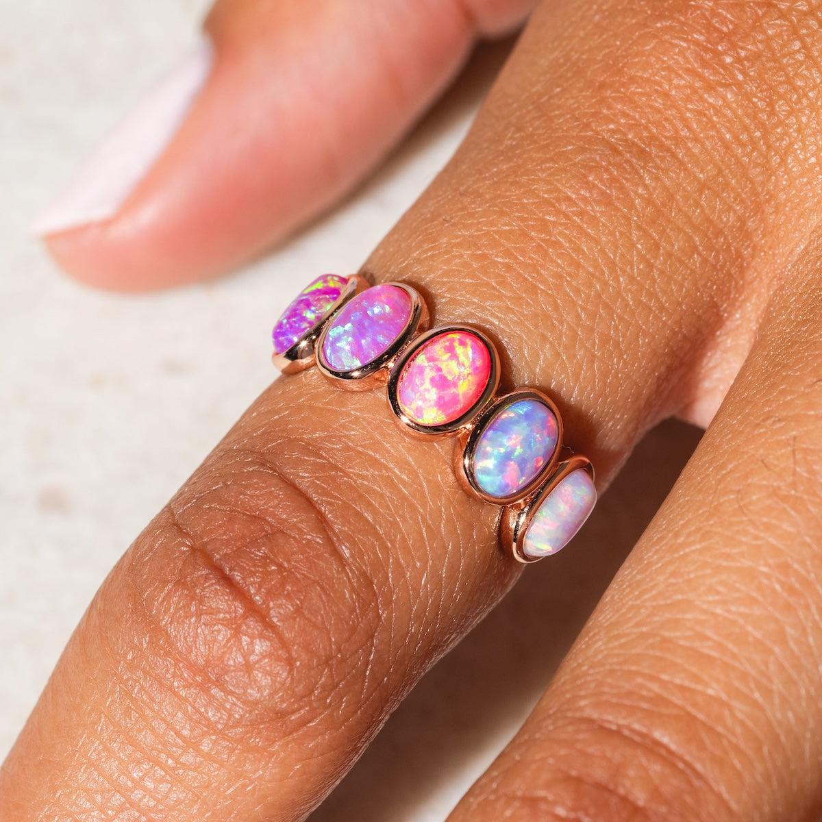 A close-up of a hand wearing the Rose Gold Opal Ombre Candy Band, featuring five oval opals in vibrant pink, purple, and blue shades, displayed across two fingers against a light background.