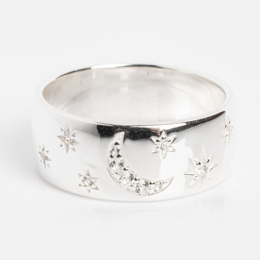 The Silver Night Sky Cigar Band is a wide rhodium plated sterling silver ring featuring engraved crescent moons and stars, accented with sparkling white topaz stones, photographed on a plain white background.