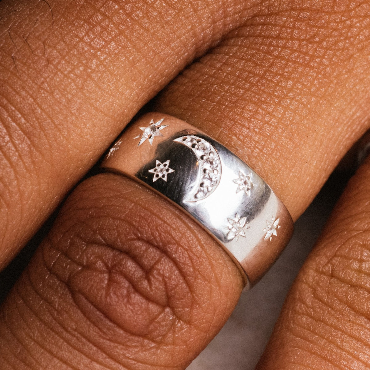 A close-up of the Silver Night Sky Cigar Band, crafted in rhodium-plated sterling silver with engraved stars, a crescent moon, and small white topaz gemstones, shown on a finger against brown skin.